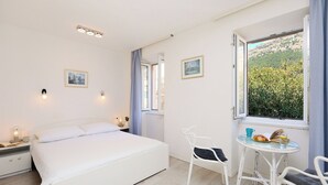 A bedroom with a bed, bedside tables, a chair, a small table with a book and glass, and a window with a view of greenery. - Sophie 1 - Old Town Heritage Suite (Dubrovnik)