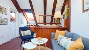 Une pièce chaleureuse avec un canapé bleu, une petite table ronde sur laquelle repose un magazine, un fauteuil bleu, des poutres en bois au plafond et un comptoir en bois avec une lampe.