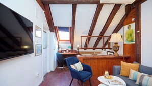 Une pièce avec un plafond en bois, un téléviseur à écran plat fixé au mur, un fauteuil bleu, un canapé avec des coussins, une petite table ronde avec un verre d’eau et un tableau accroché au mur.