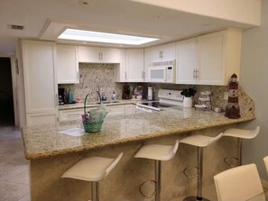 A modern kitchen with white cabinets, a granite countertop, and a bar area with stools. - SpaceX Launches and Fun Filled Family Beach Days - 1st Floor 2BR/2BA Condo (South Padre Island)