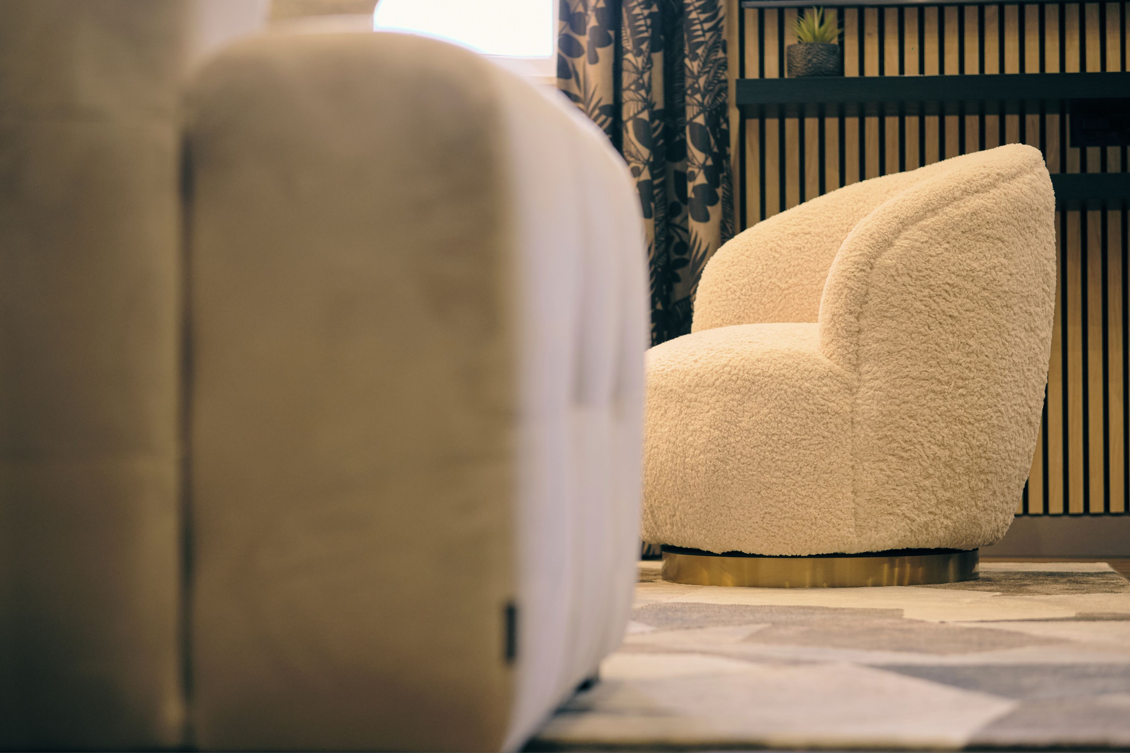 Een comfortabele fauteuil met een ronde basis, geplaatst voor een patroonrijk gordijn en een plank met een plant.