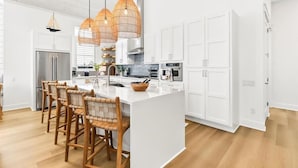 A modern kitchen with white cabinetry, a large island, wicker chairs, and pendant lights. - Sol Seekers 109 PLA (Port Aransas)