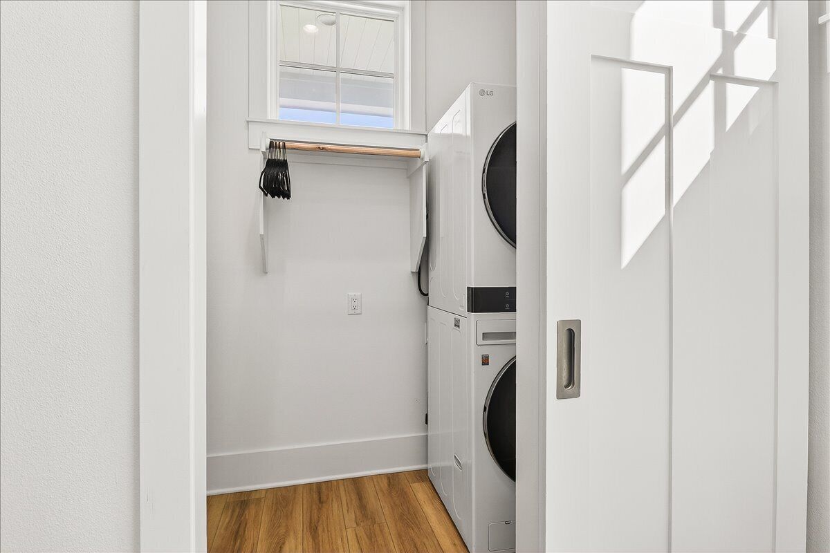 A laundry room with two stacked washers and dryers, a wooden floor, a white wall, and a window above the washing machine.