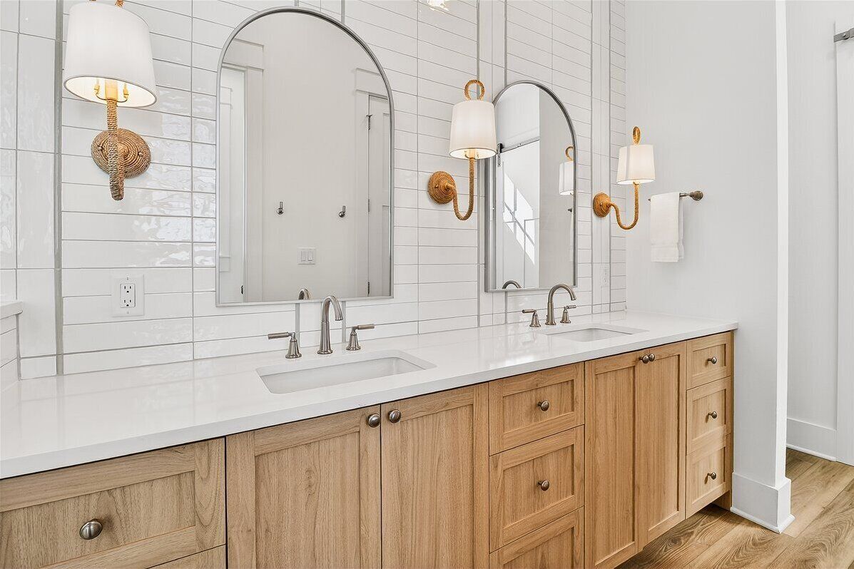 Un baño con pared de azulejos blancos, tres espejos, dos lavamanos, armarios de madera y apliques de pared.