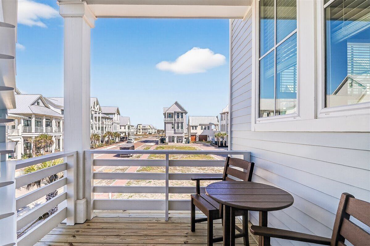 A balcony with a view of a coastal street, wooden deck, and a table with chairs.
