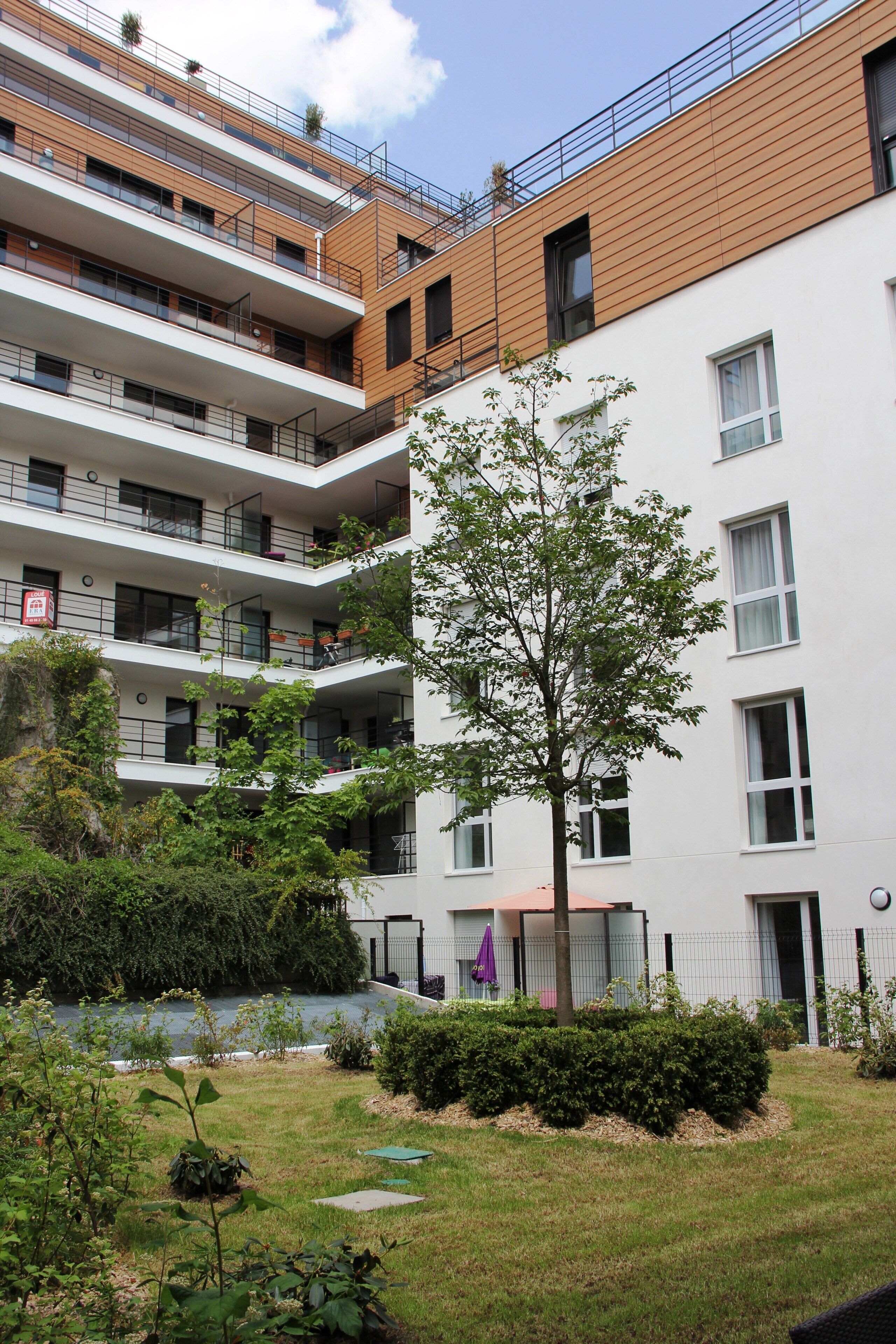 Un immeuble résidentiel à plusieurs étages, doté de balcons, avec un arbre dans la cour et un petit jardin.