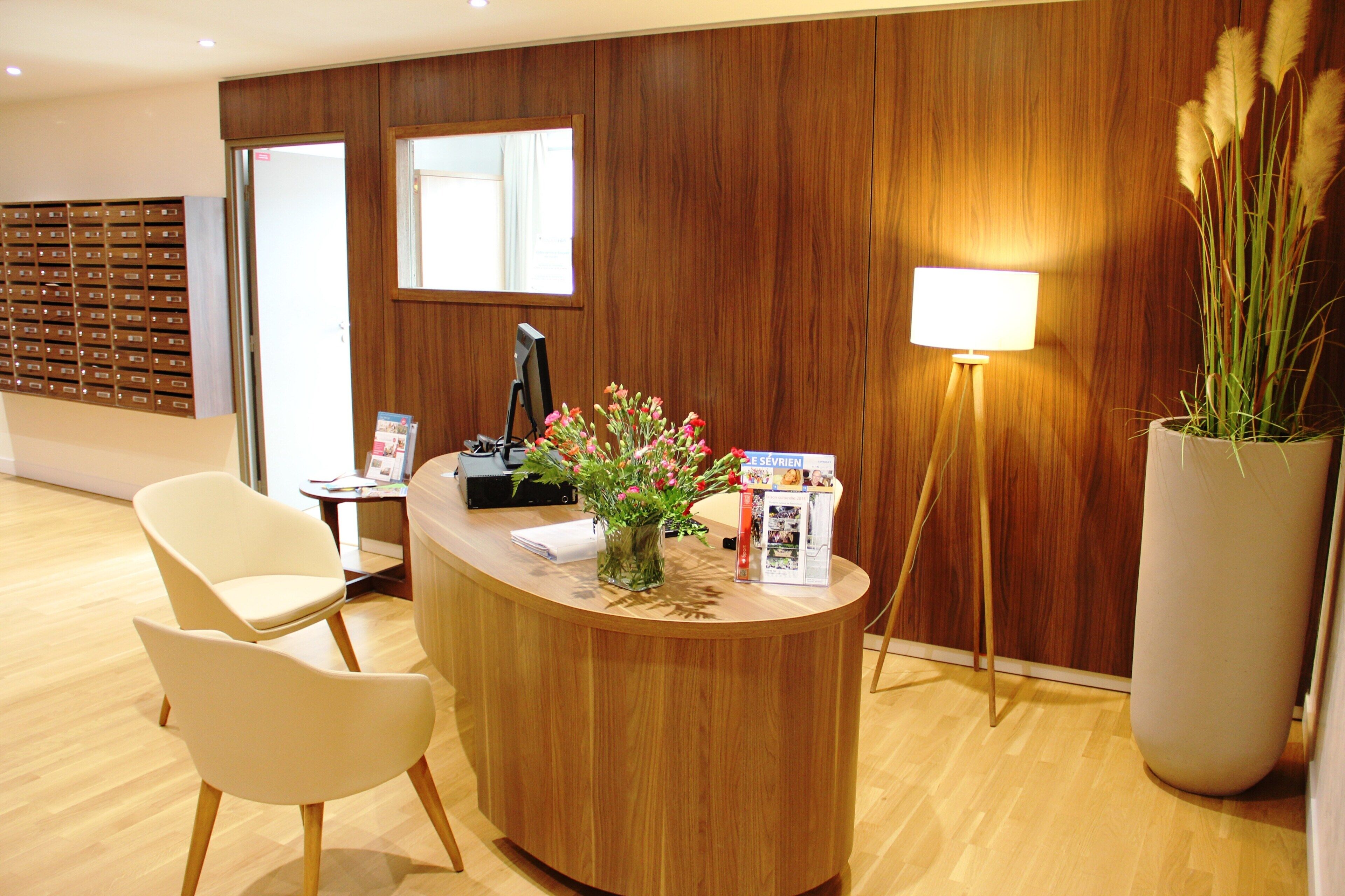 Un comptoir d’accueil avec un revêtement en bois, une chaise blanche, une lampe de bureau, un grand vase rempli de fleurs et un panneau d’affichage avec des fentes pour le courrier.