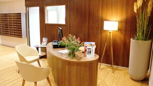 En receptionsdisk med träinredning, en vit stol, en skrivbordslampa, en stor vas med blommor och en anslagstavla med fack för post.