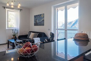 A living room with a sofa, a dining table, a fruit bowl, a painting, and a view of mountains through large windows. - Alba on Lake Como (Sala Comacina)