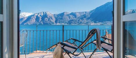 A balcony with a chair and a woven basket, overlooking a body of water and mountains.