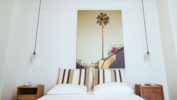 A hotel room with a bed, a nightstand, a wall-mounted painting of a palm tree, and two pendant lights. - we saw athens from 4b (Athina)