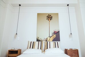 A hotel room with a bed, a nightstand, a wall-mounted painting of a palm tree, and two pendant lights. - we saw athens from 4b (Athina)