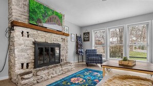 A living room with a stone fireplace, a blue floral rug, a blue armchair, and a window with blinds. - Lakeside Pines: Waterfront > Bike in/out > Game Room > Fitness Room (Bella Vista)