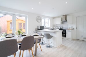 A modern kitchen with white cabinetry, a central island, and a dining area with grey chairs and a table set for two. - Lytham St Annes FY8 | 3BR Home | Near Blackpool (Lancashire)