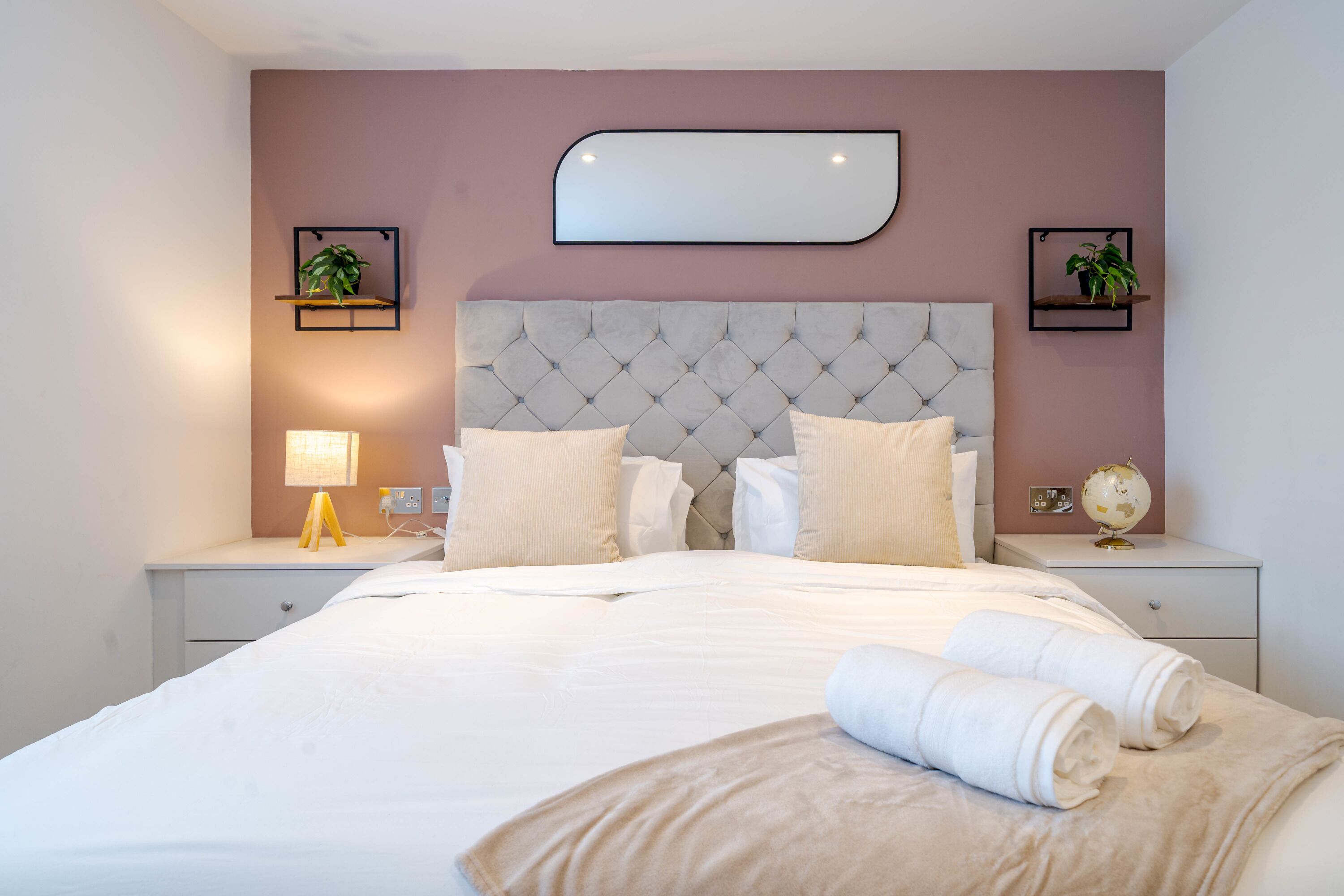 A neatly made bed with a tufted headboard, two bedside tables with lamps, and two wall-mounted shelves with plants.