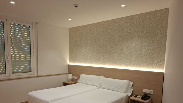 Une chambre d’hôtel avec deux lits, une tête de lit dotée d’un éclairage intégré, une table de chevet avec un téléphone et une fenêtre avec des stores.