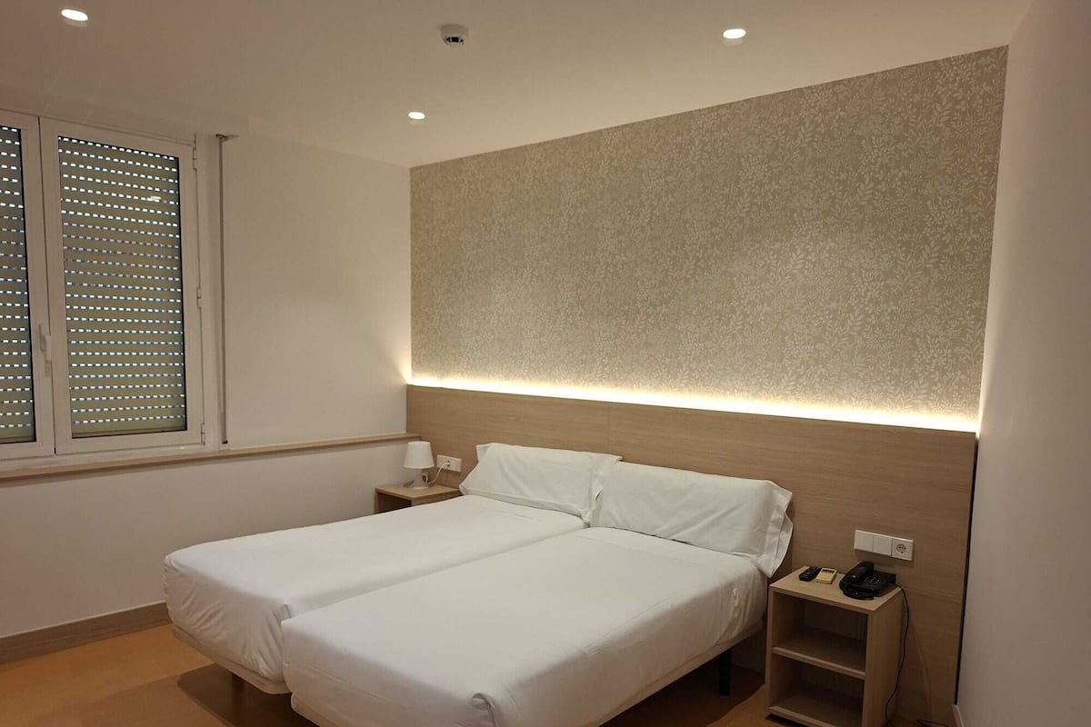 Une chambre d’hôtel avec deux lits, une tête de lit dotée d’un éclairage intégré, une table de chevet avec un téléphone et une fenêtre avec des stores.