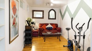Uma sala de estar com um sofá vermelho, uma mesa de centro de vidro, uma bicicleta ergométrica e um espelho fixado na parede.