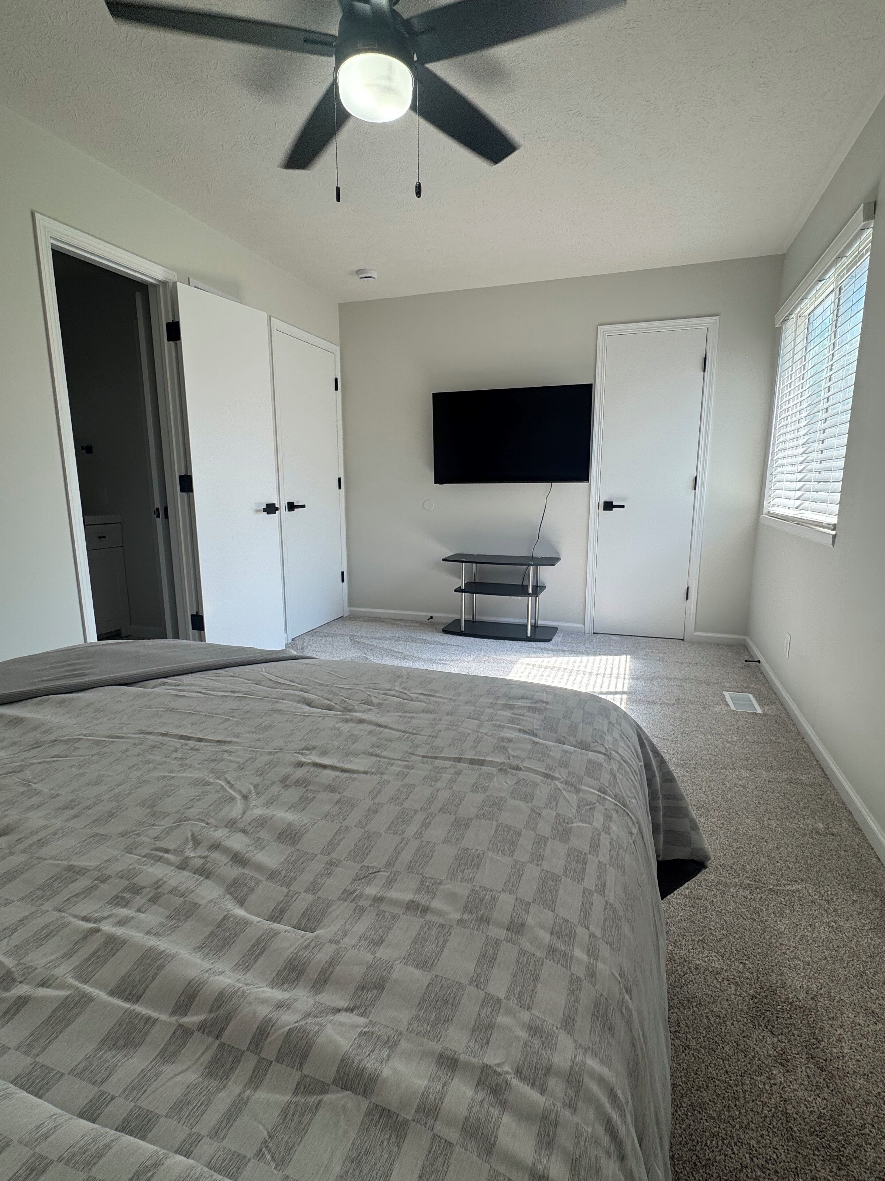Um quarto com cama, ventilador de teto, televisão fixada na parede e janela com persianas.