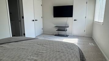 Um quarto com cama, ventilador de teto, televisão fixada na parede e janela com persianas.