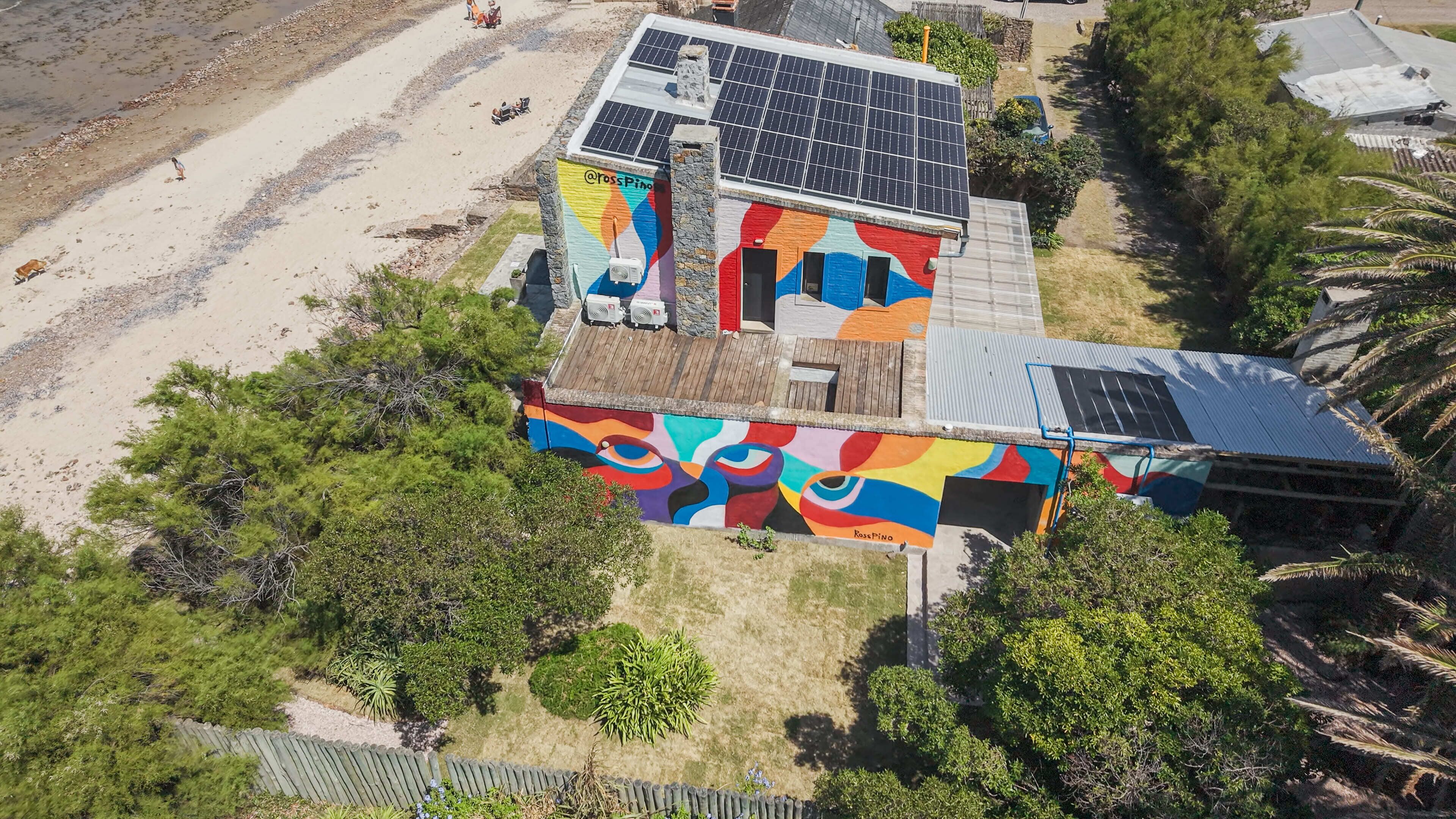 En ejendom ved stranden med et farverigt muralmaleri, solpaneler og en indhegnet have.