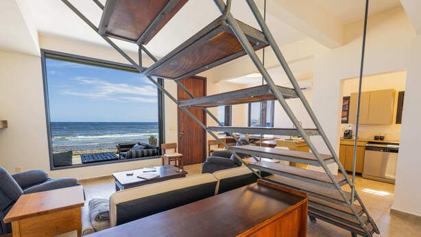 A modern interior with a staircase leading to a balcony overlooking the ocean, a dining area, and a kitchen. - Direct Beach Access!! - Casa On The Rocks Uruguay (Estación Las Flores)