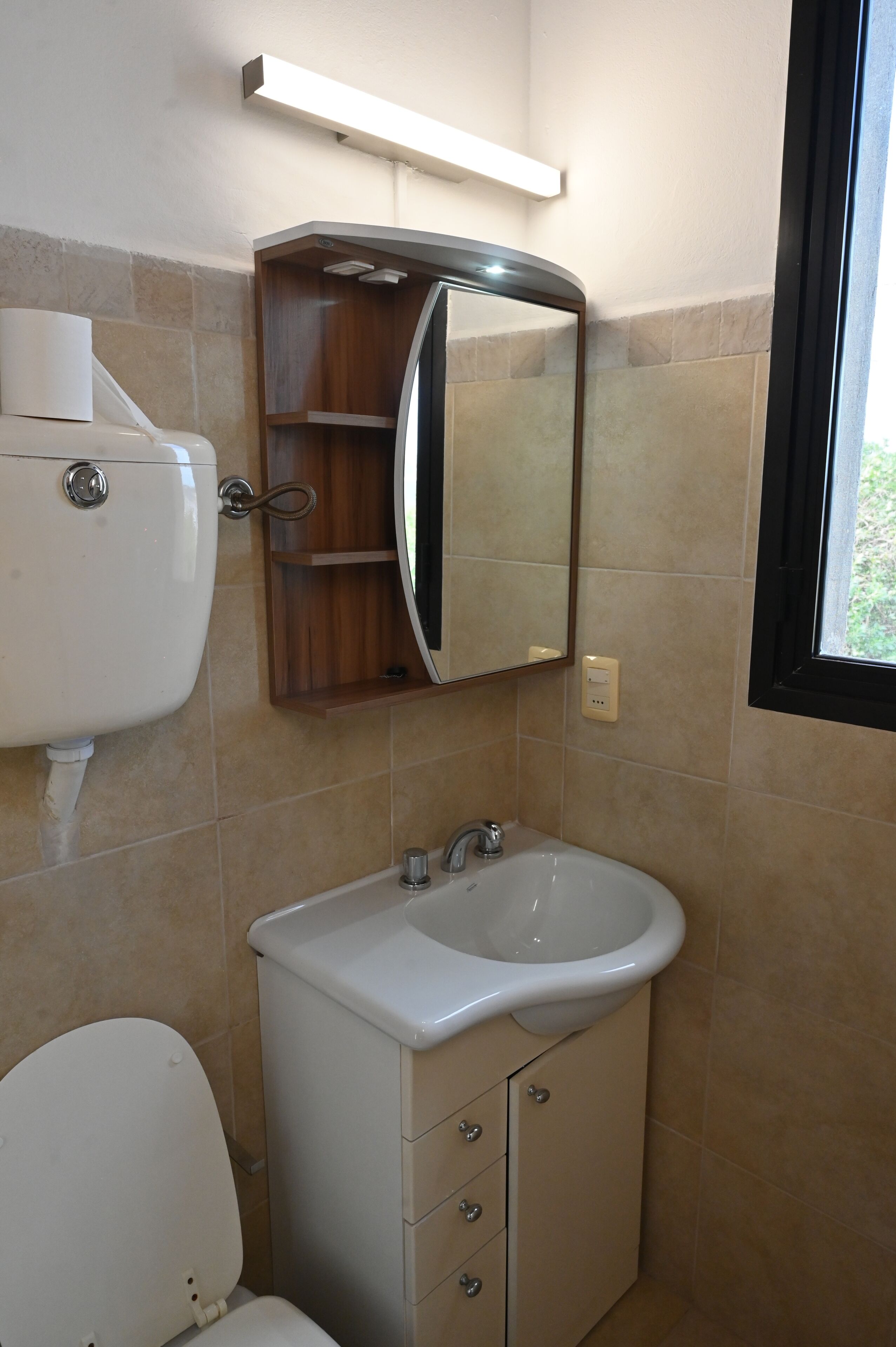 Et badeværelse med toilet, vask, spejl og et vindue.