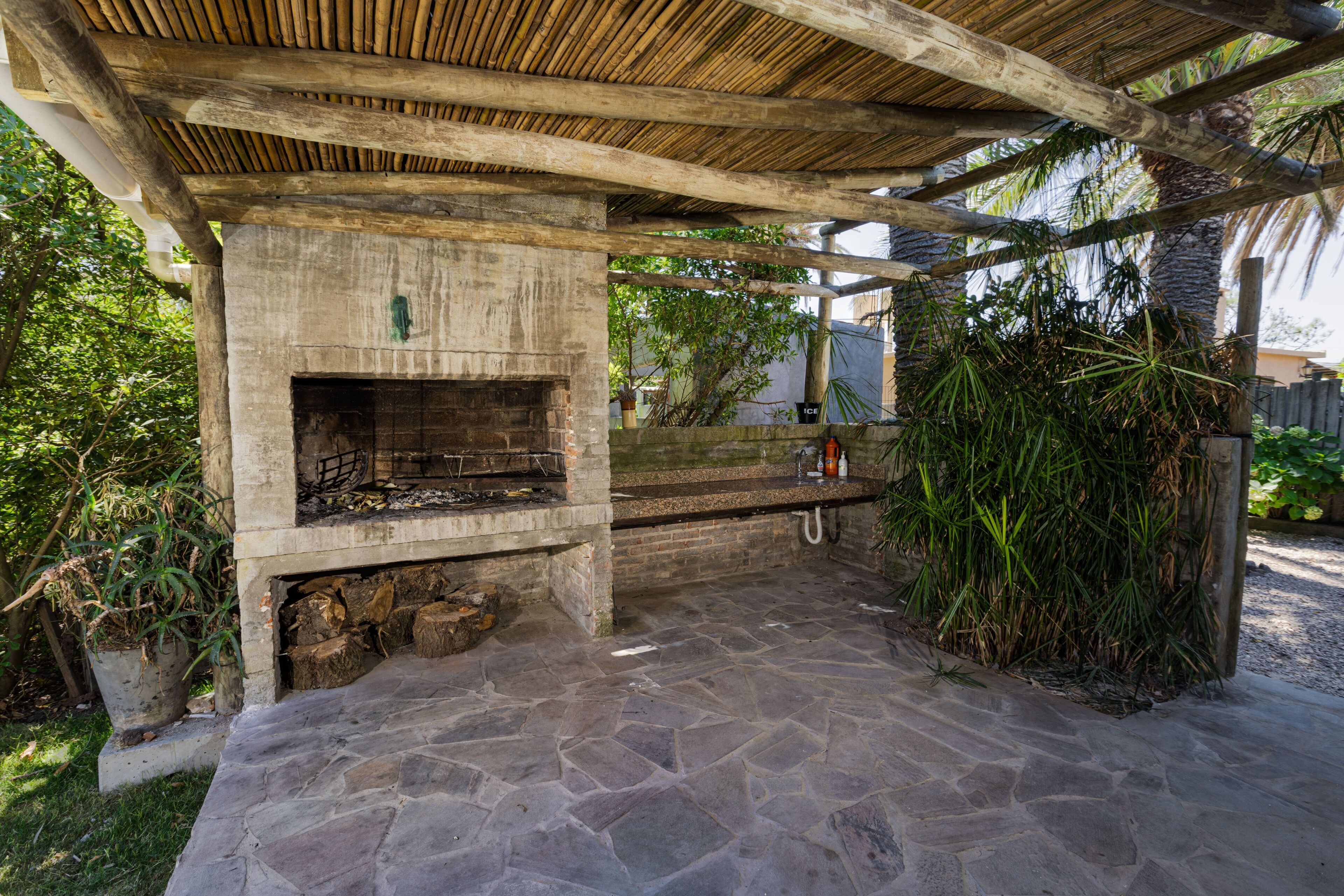 A stone patio with a built-in grill area, surrounded by greenery and a wooden pergola structure.