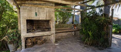 A stone patio with a built-in grill area, surrounded by greenery and a wooden pergola structure.