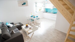 A modern living room with a sofa, a small table, and a dining area with chairs. A staircase is visible in the background. - Baltic Stone - Comfortable vacation homes in Rewal (Rewal)