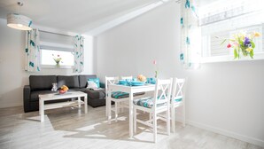 A modern living room with a white sofa, a dining table with chairs, and a window with curtains. - Baltic Stone - Comfortable vacation homes in Rewal (Rewal)