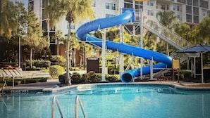 Una piscina con un tobogán de agua azul, rodeada por un edificio de hotel y palmeras.