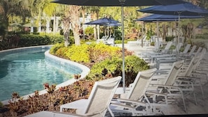 Zona junto a la piscina con sillones de descanso y sombrillas, rodeada de vegetación y árboles.