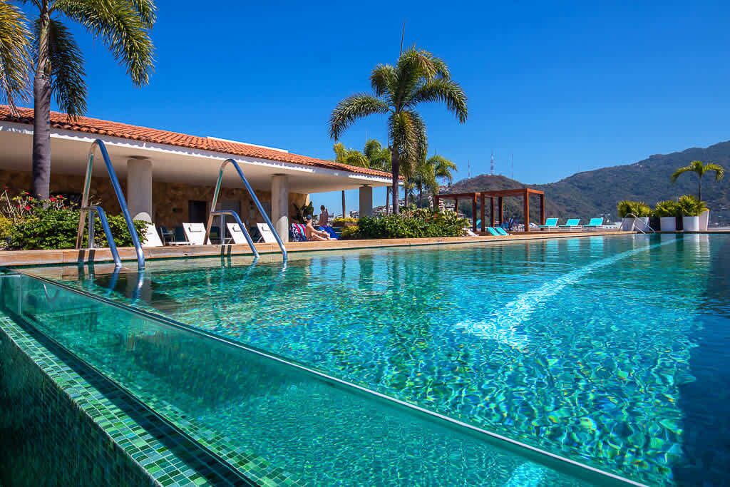 Una piscina cristalina con sillas de descanso, palmeras y un edificio con techo de tejas.