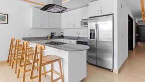 Eine moderne Küche mit weißen Schränken, einem Edelstahlkühlschrank, einer integrierten Kochinsel mit Barhocker und einer Kaffeemaschine.