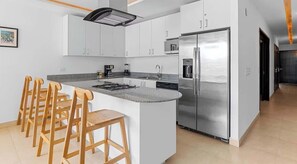 A modern kitchen with white cabinets, a stainless steel refrigerator, a built-in island with bar stools, and a coffee machine. - The Park 306 | Ocean View • Heated Pool • (Puerto Vallarta)