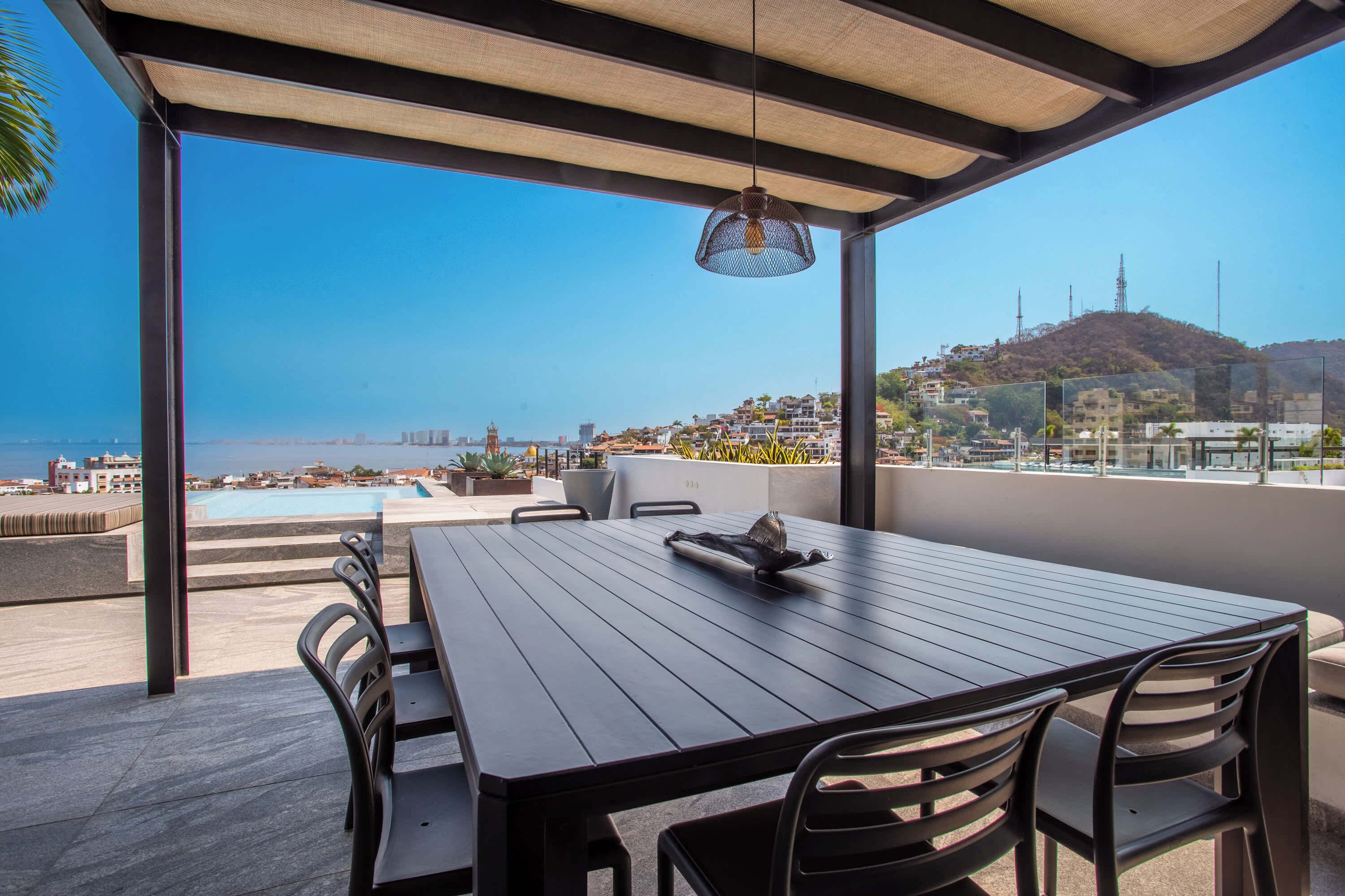 A covered outdoor seating area with a table and chairs, overlooking a pool and cityscape.