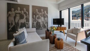 Ein modernes Wohnzimmer mit einer Couch, Couchtisch und Fernseher. An den Wänden hängen zwei große Poster und vom Balkon hat man einen Blick auf die Stadt.