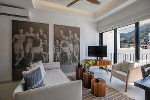 Ein modernes Wohnzimmer mit einer Couch, Couchtisch und Fernseher. An den Wänden hängen zwei große Poster und vom Balkon hat man einen Blick auf die Stadt.