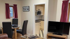 Un soggiorno arredato con divano, tavolino in vetro, televisione, angolo da pranzo con sedie e cucina visibile attraverso una porta aperta.