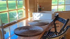 Un intérieur en bois avec une table à manger, deux sets de table en fibres tressées, une chaise noire et une fenêtre donnant sur un espace verdoyant.
