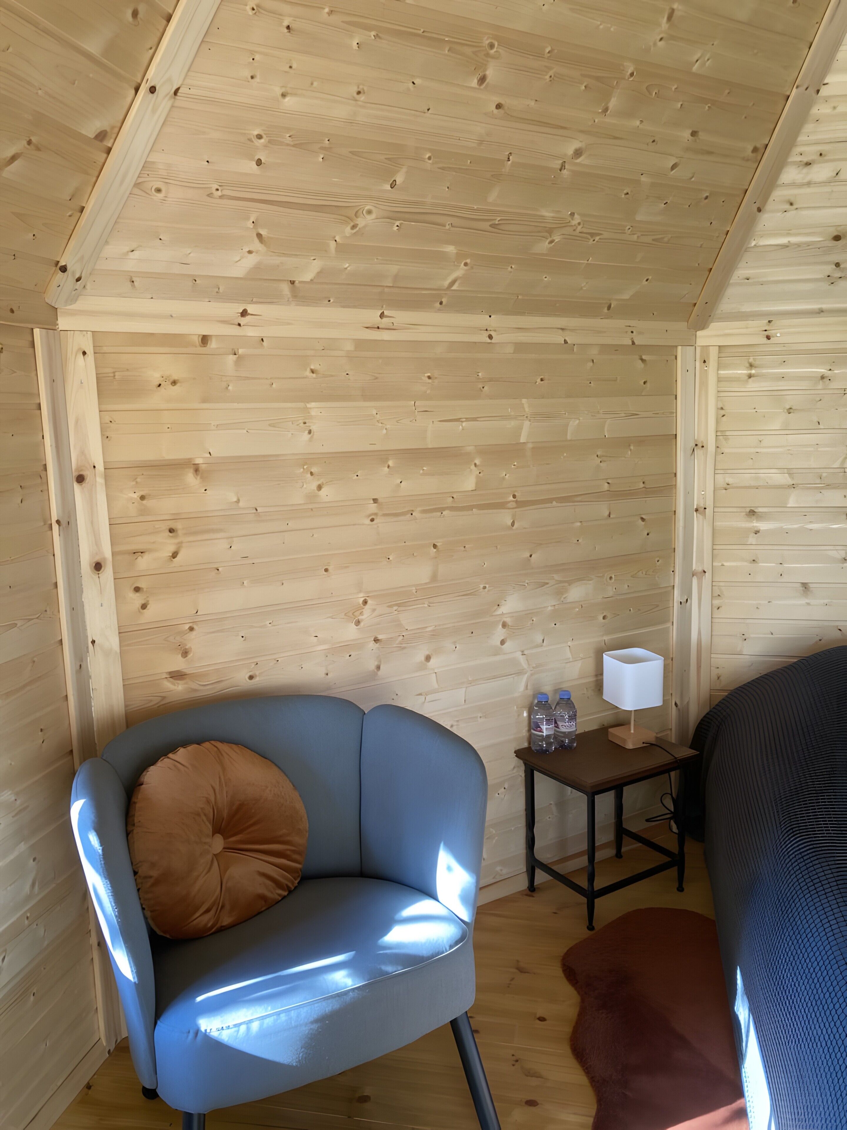 Une petite pièce chaleureuse avec un plafond en bois, une chaise bleue avec un coussin marron, une petite table avec une lampe et des bouteilles d’eau, et un lit recouvert d’une couverture à motifs.