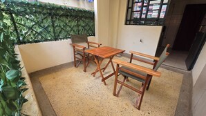 Een klein terras buiten met een tafel, stoelen, een groene plantenwand en een raam met uitzicht op gebouwen.