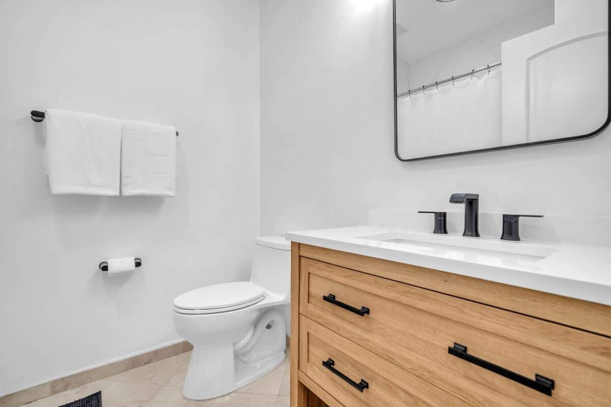 A modern bathroom with a toilet, a white sink with black handles, a wooden vanity, and a mirror above the sink.