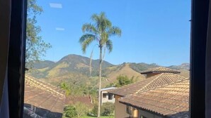 A window view of a rural landscape with hills, a palm tree, and a house with a tiled roof. - Villa Mantiqueira SFX - Suítes (São José dos Campos)