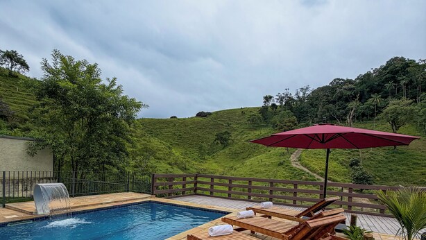 A swimming pool with wooden lounge chairs and a red umbrella, surrounded by a stone deck and lush greenery. - Villa Mantiqueira SFX - Suítes (São José dos Campos)