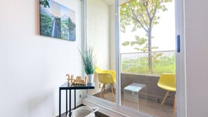 Un balcon avec une porte vitrée, une petite table ornée d’une plante, deux chaises jaunes et une pancarte de bienvenue.
