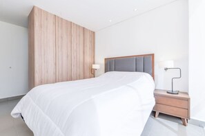 Un dormitorio moderno con una cama grande, un armario de madera, una mesita de noche con una lámpara y una mesita auxiliar.