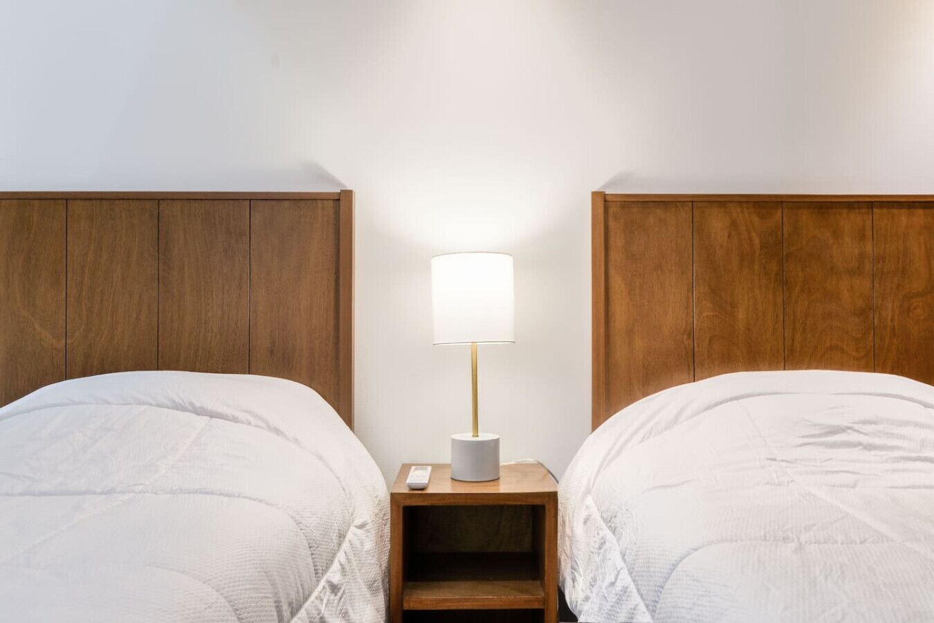 A hotel room with two beds, a nightstand with a lamp, and a remote control.