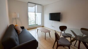 Un salon moderne comprenant un canapé, une table en verre et des chaises, un téléviseur à écran plat fixé au mur et une grande fenêtre laissant entrer la lumière naturelle.